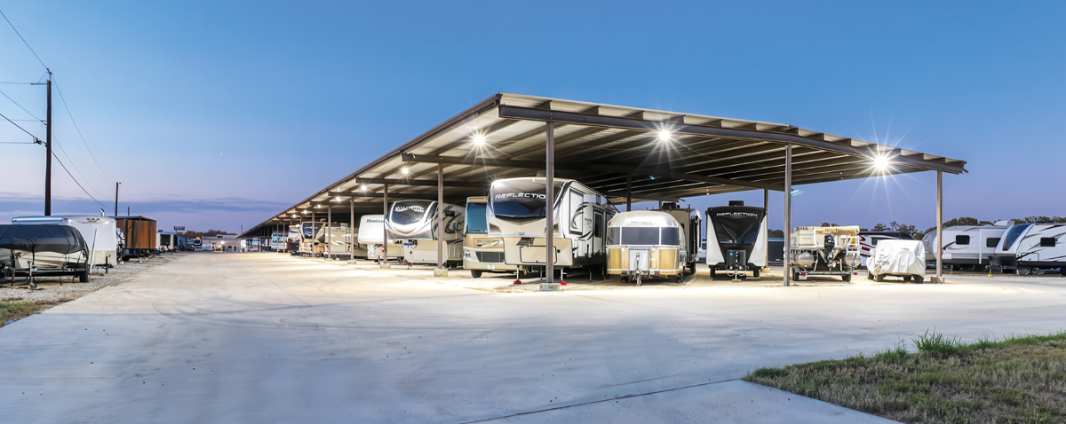 RVs parked under a lit canopy at twilight in a large concrete area.