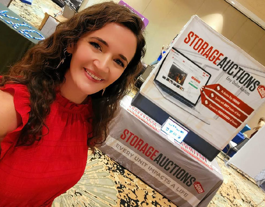 Alonna Ross smiling at an industry trade show, standing next to a 'StorageAuctions.com' booth display promoting online auctions.