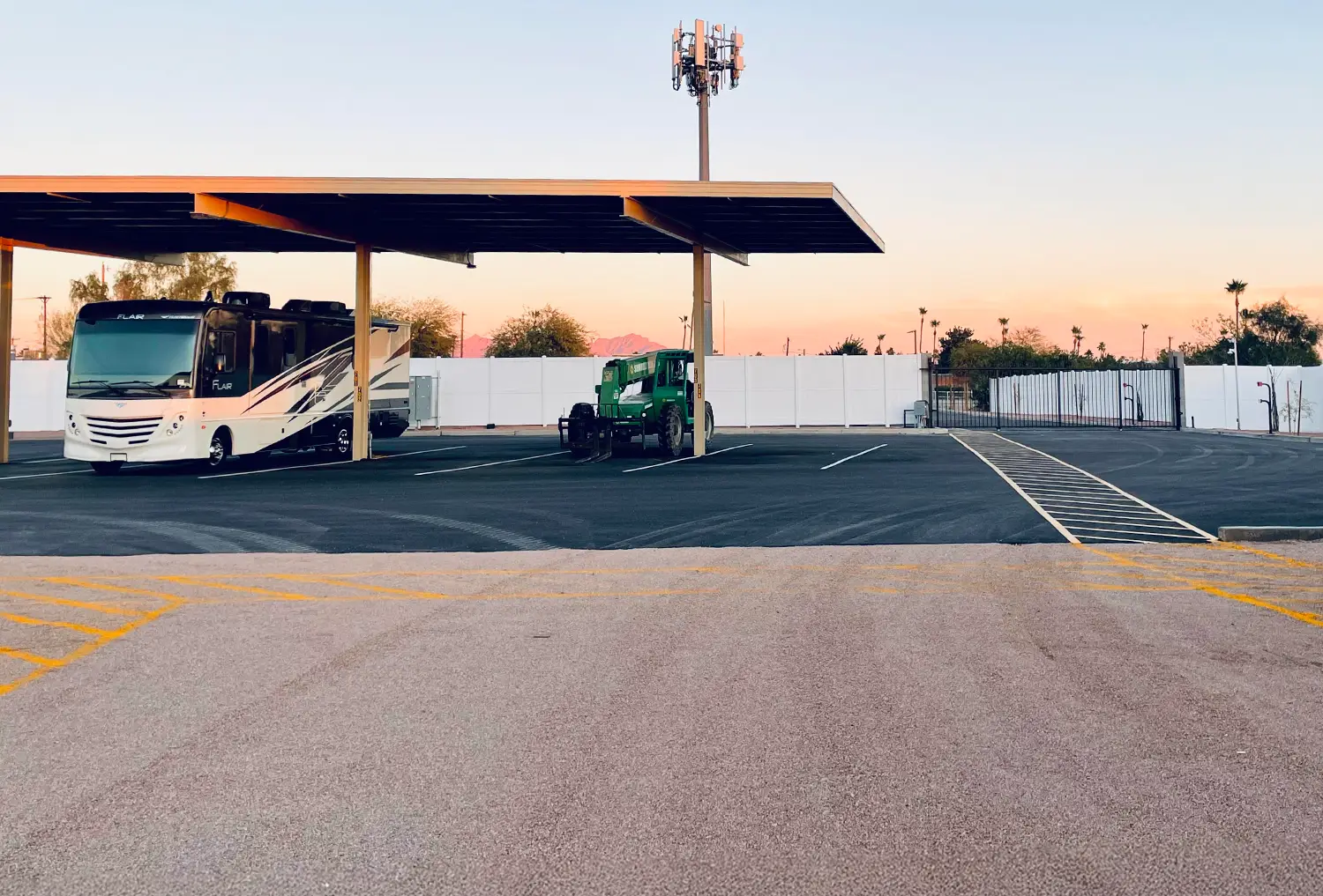 RV and equipment parked under a covered storage area at an outdoor storage facility, with a cell tower and sunset sky in the background.