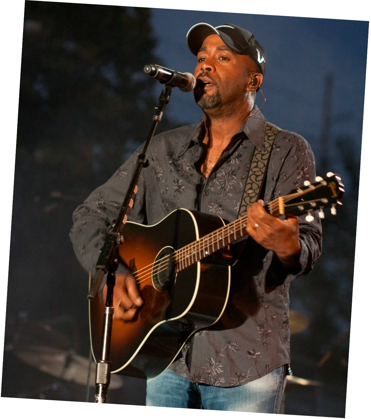 Darius Rucker, wearing a baseball cap and patterned shirt, singing and playing guitar.