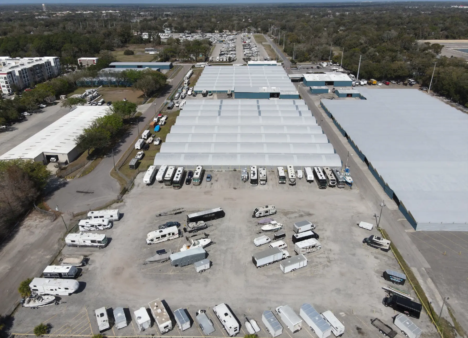 Landscape orientation aerial photograph view of a large outdoor RV and boat storage facility space with numerous recreational vehicles (RVs) and boats parked in open lots; This pictured facility space happens to be one of Ramser Development Company's RV and boat storage facilities; Several long, low buildings with light-colored roofs are also visible, some covered, suggesting more storage units; The facility space is surrounded by trees and some residential or commercial buildings in the far distant background