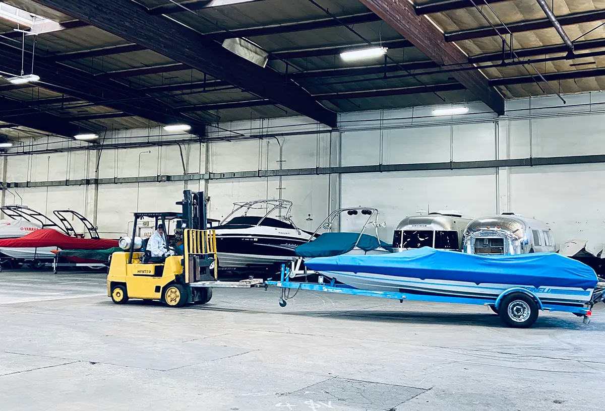 Landscape orientation interior photograph view of a large warehouse with high ceilings and exposed beams; A yellow forklift with person operating it is moving a boat that is covered with a blue tarp and attached to a trailer; Other boats are visible in the background, stored within the facility; The pictured boat happens to be towed at one of Ramser Development Company's RV and boat storage facilities