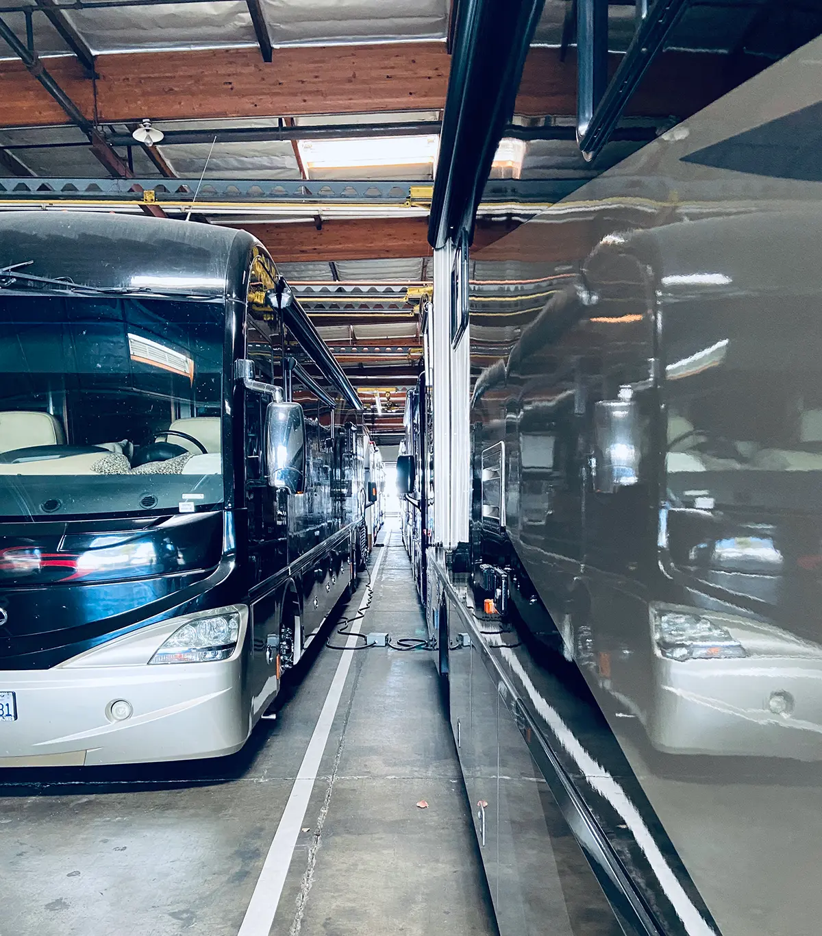 Portrait orientation interior photograph view of a narrow aisle between two large, dark-colored recreational vehicles (RVs) or motorhomes parked indoors, likely in a storage facility; The front of the RV on the left is visible, showing its headlights and part of the windshield; The roof structure of the building is visible above; This picture happens to show RVs in their respective charging phase at one of Ramser Development Company's RV and boat storage facilities
