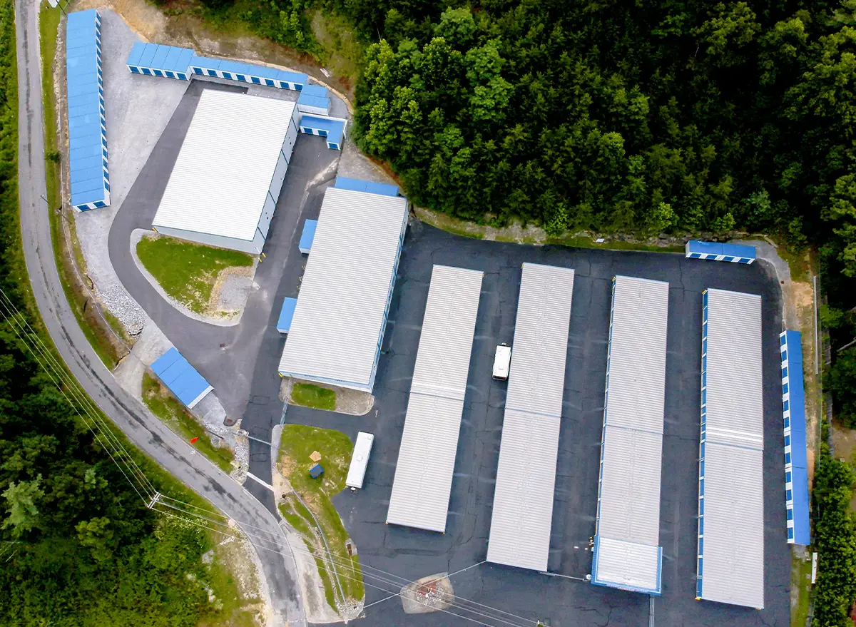 Aerial drone view of a modern self-storage facility with multiple long white buildings and blue-doored units, surrounded by trees and a paved driveway.