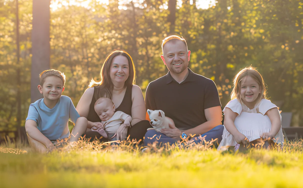 A smiling family of four with a baby and a small white dog, sitting together in a grassy field with golden sunlight in the background.