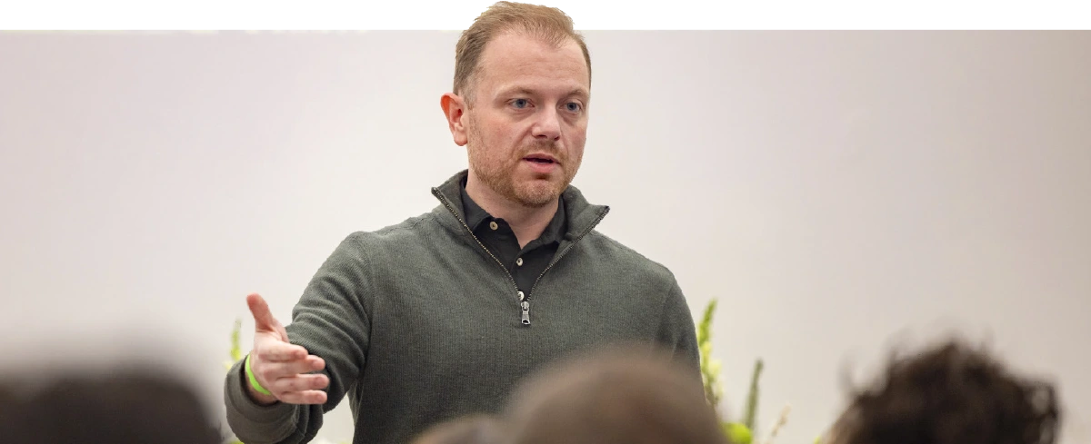 A man with a beard wearing a green sweater speaking to an audience and gesturing with his hand in a professional setting.