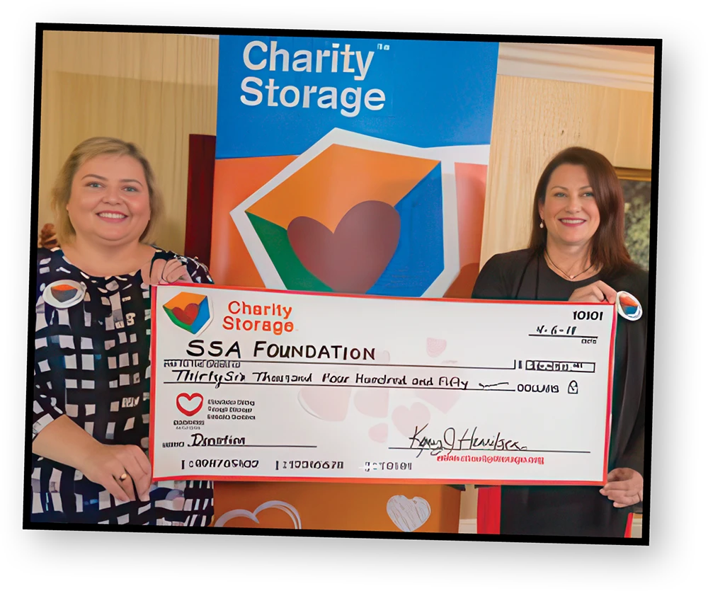 Two women smile as they hold up a large novelty check made out to the "SSA Foundation" for thirty-six thousand four hundred and fifty dollars. A banner with the "Charity Storage" logo is visible in the background.
