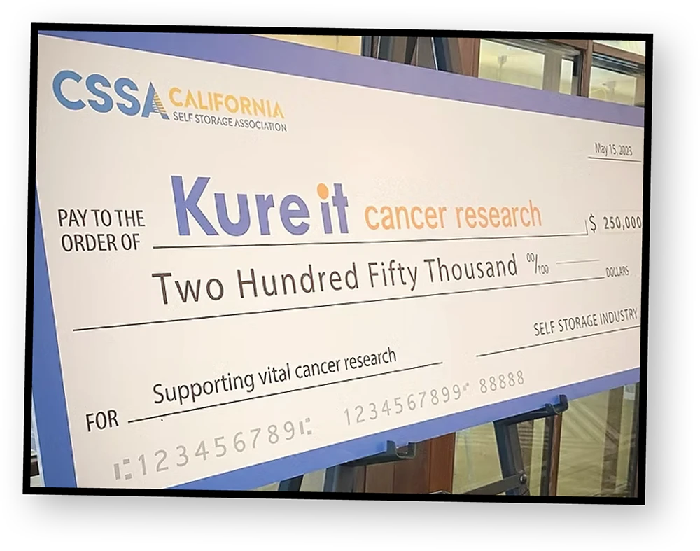 A close-up shot of a large novelty check on a stand, made out to "Kureit cancer research" for two hundred and fifty thousand dollars. The check is sponsored by the "California Self Storage Association."