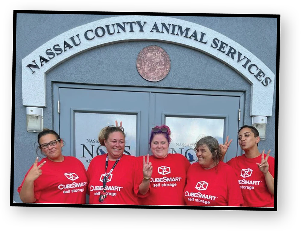 A group of five women wearing matching red shirts with the CubeSmart logo pose for a photo in front of a building entrance with "Nassau County Animal Services" written above the door. They are all smiling and holding up a peace sign with their hands.