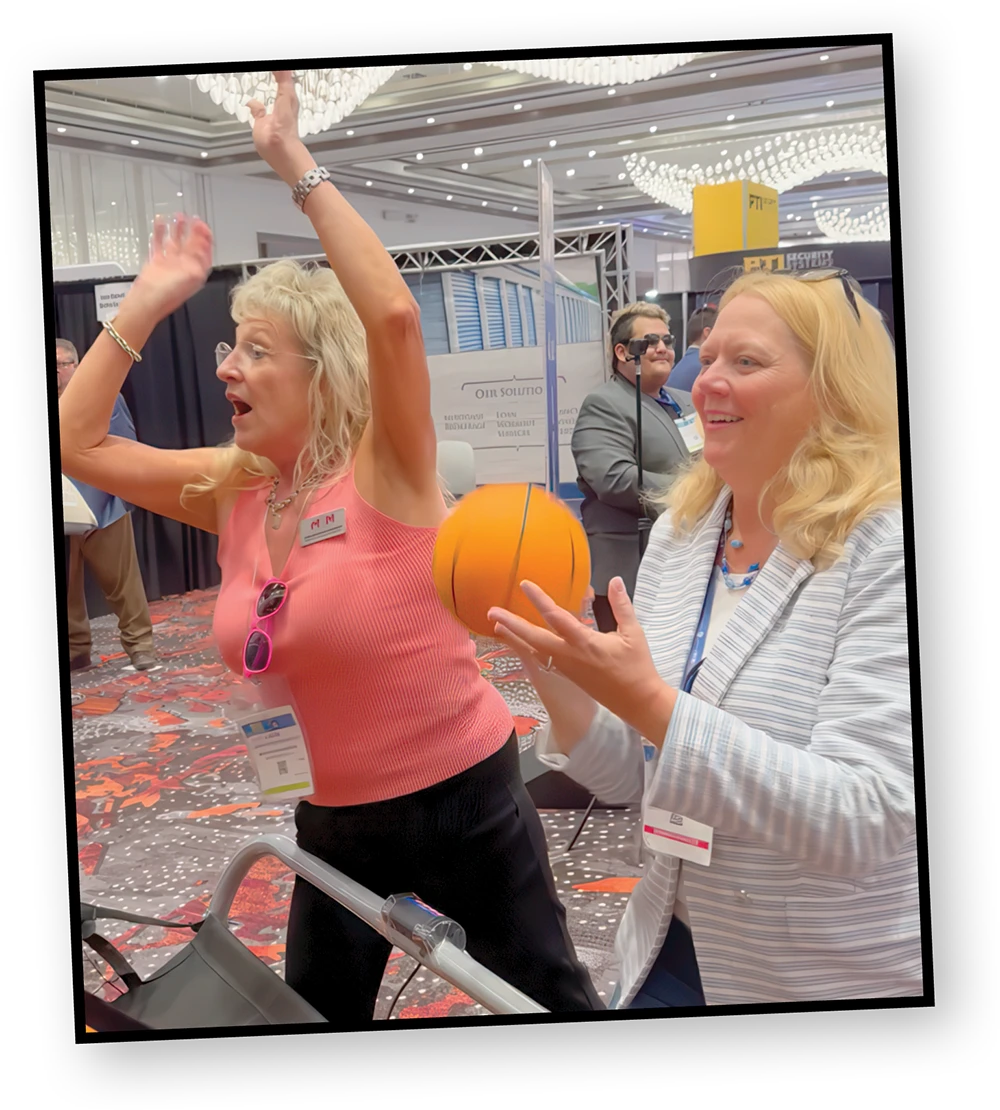 Two women play a game at a conference. One woman on the left in a pink shirt is throwing an orange basketball while the woman on the right in a striped blazer is preparing to shoot it.