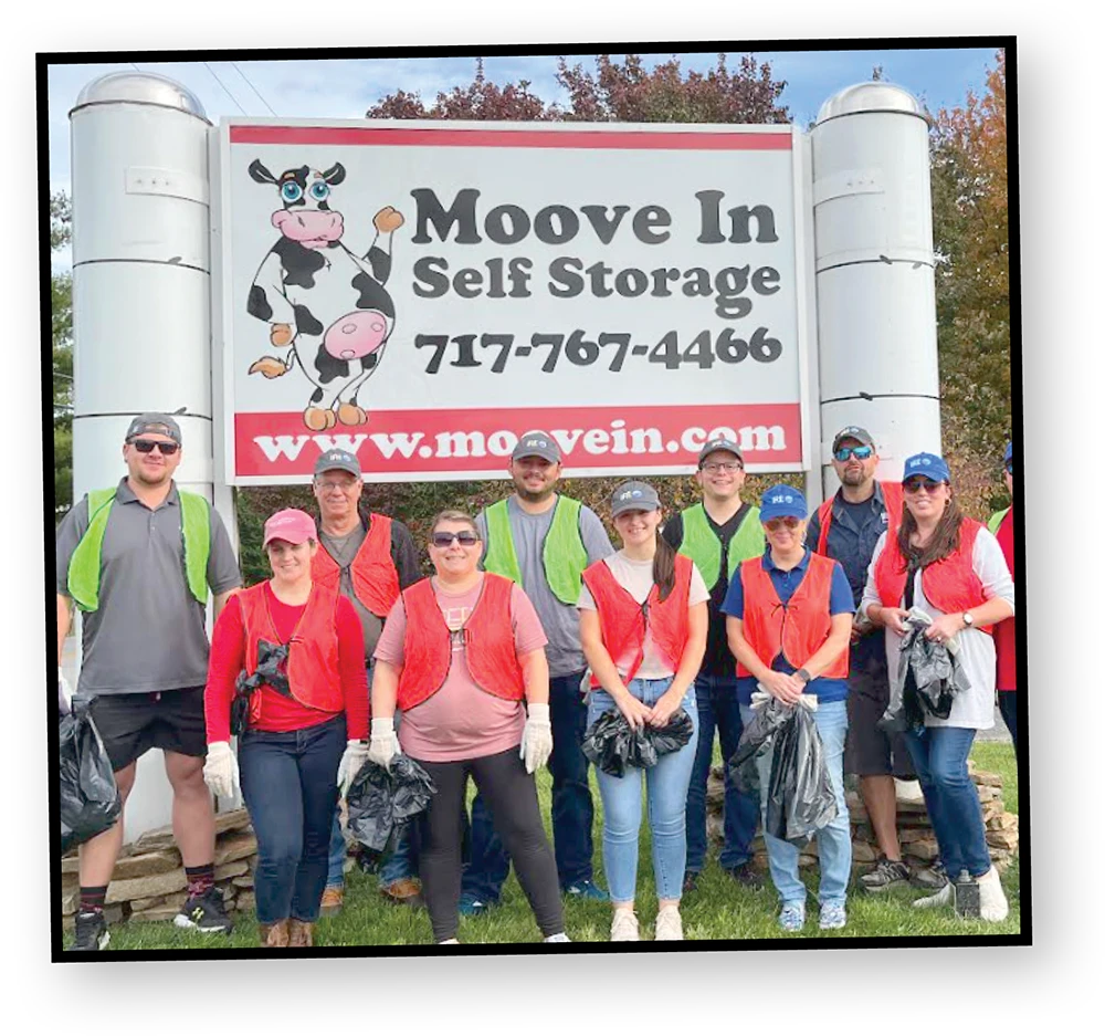 A group of people stand together for a photo in front of a "Moove In Self Storage" sign with a cow mascot on it. Most of the people are wearing bright orange or neon green vests and holding black garbage bags.