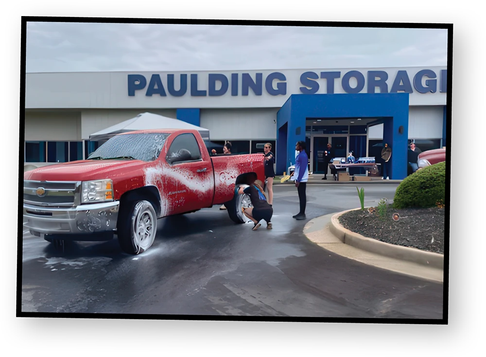 A red truck is being washed in front of a building that says, "Paulding Storage" in large blue letters. One person is hosing the truck while another is bending down to scrub a tire.