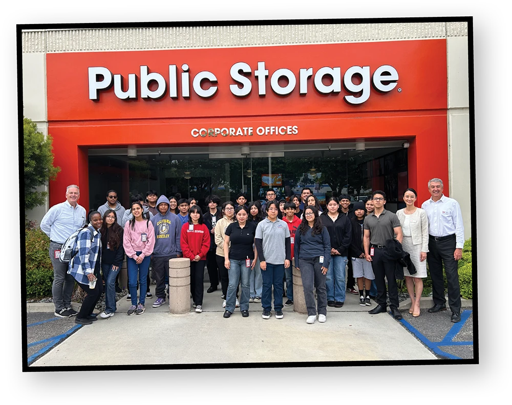 A large group of students and a few adults pose for a group photo in front of the Public Storage corporate office building. The sign above the door reads, "Public Storage" in large red letters.