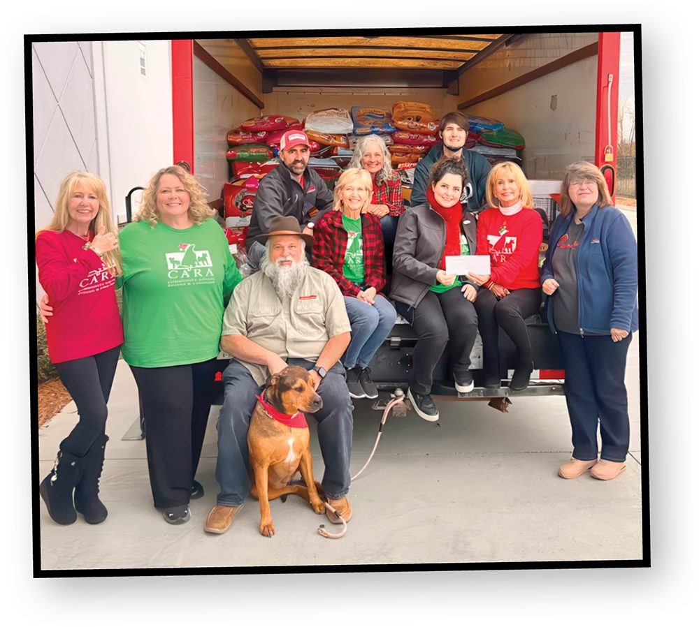 A group of people and one dog pose for a photo in front of a large truck with the back open, revealing bags of what looks like dog food. The people are smiling, and one man is sitting with the dog on his lap.