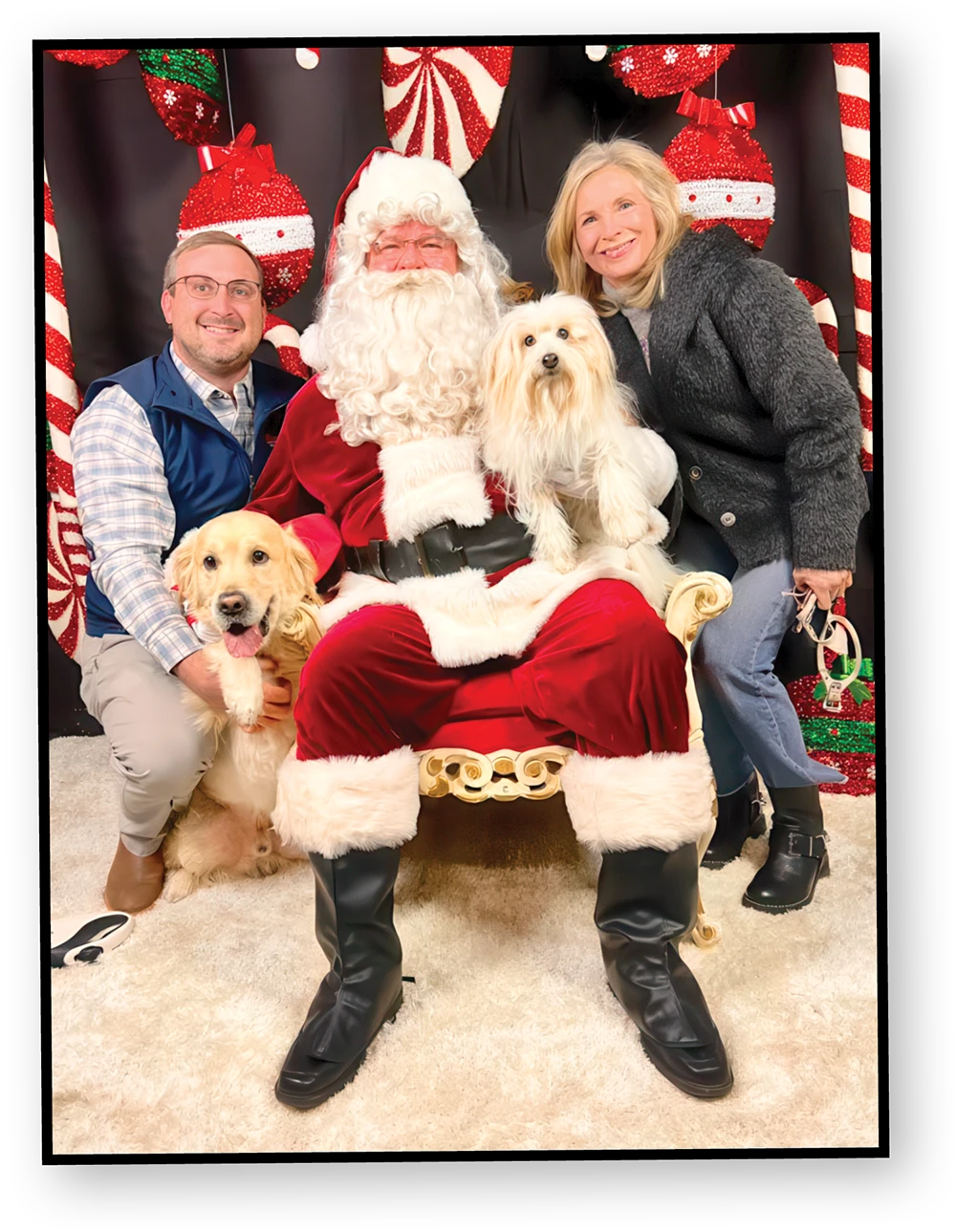 A man and a woman pose with Santa Claus. The woman is sitting on the arm of his chair while the man is kneeling beside him. Santa is holding a small white dog and a golden retriever is sitting on the floor beside him.
