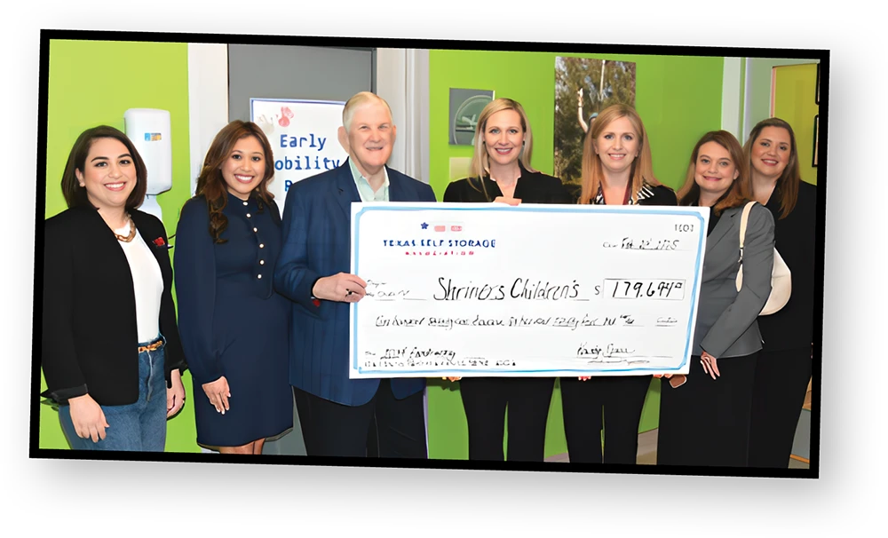 A group of six people stand together, holding a large check made out to "Shriner's Children's" for $179,644. The people are all smiling at the camera and standing in front of a lime green wall.