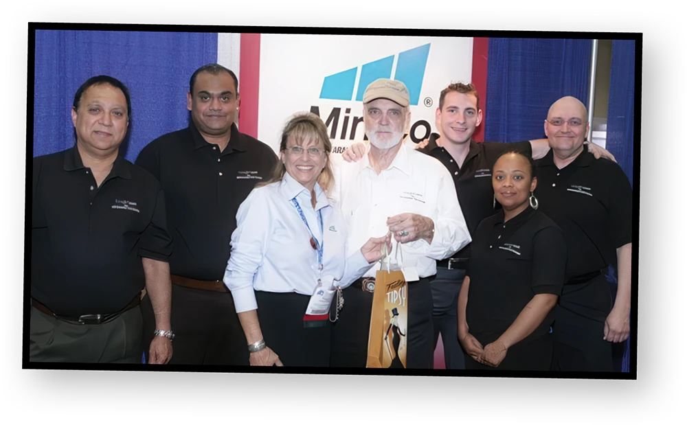 A group of six people wearing black or light blue shirts with the "Miro" logo on them are smiling and looking at the camera. An older man in the middle with a beard is holding a bag.