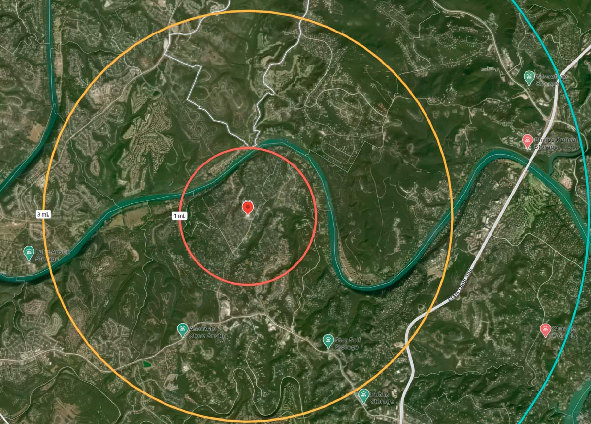 An aerial map view centered on a red map pin. There are two concentric circles around the pin. The inner red circle is labeled "1 mi." and the outer orange circle is labeled "3 mi." The area within the circles is primarily green, with a winding river and some roads and buildings visible. Several other self-storage facility pins are visible outside the circles.