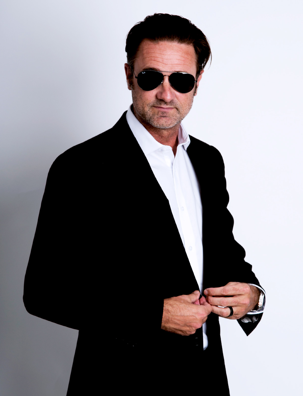 Travis Morrow wearing a black suit jacket, an open-collar white shirt, and aviator sunglasses.