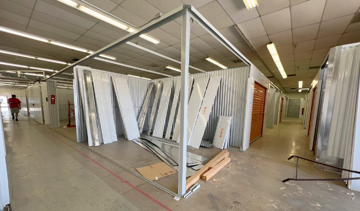 View down a hallway inside an indoor self-storage facility, showing a section of units under construction with metal wall panels leaning against a newly erected steel frame. 