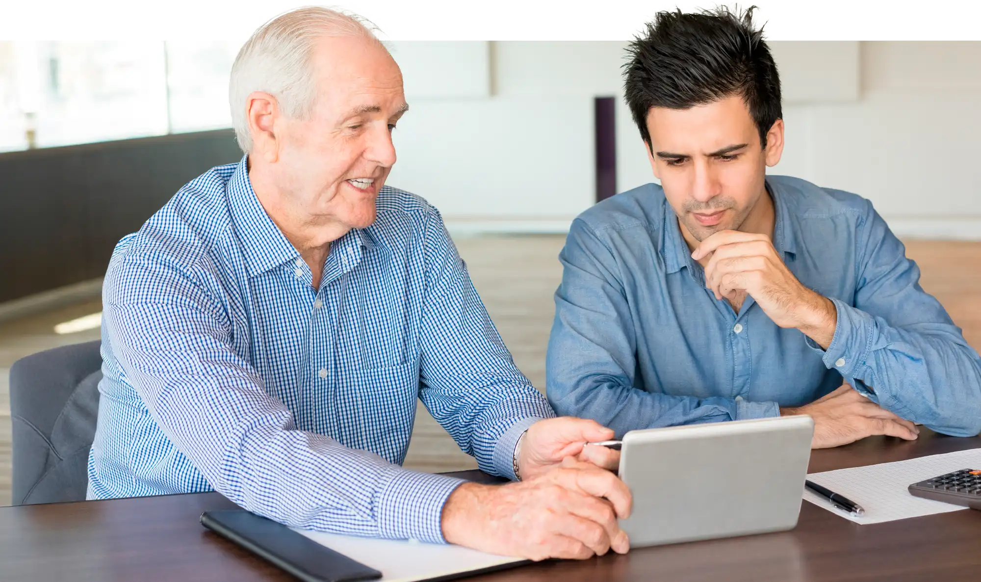 Two men, an older man with gray hair and a younger man with dark hair, are seated at a table. The older man is holding a tablet and pointing to the screen, while the younger man looks on intently.