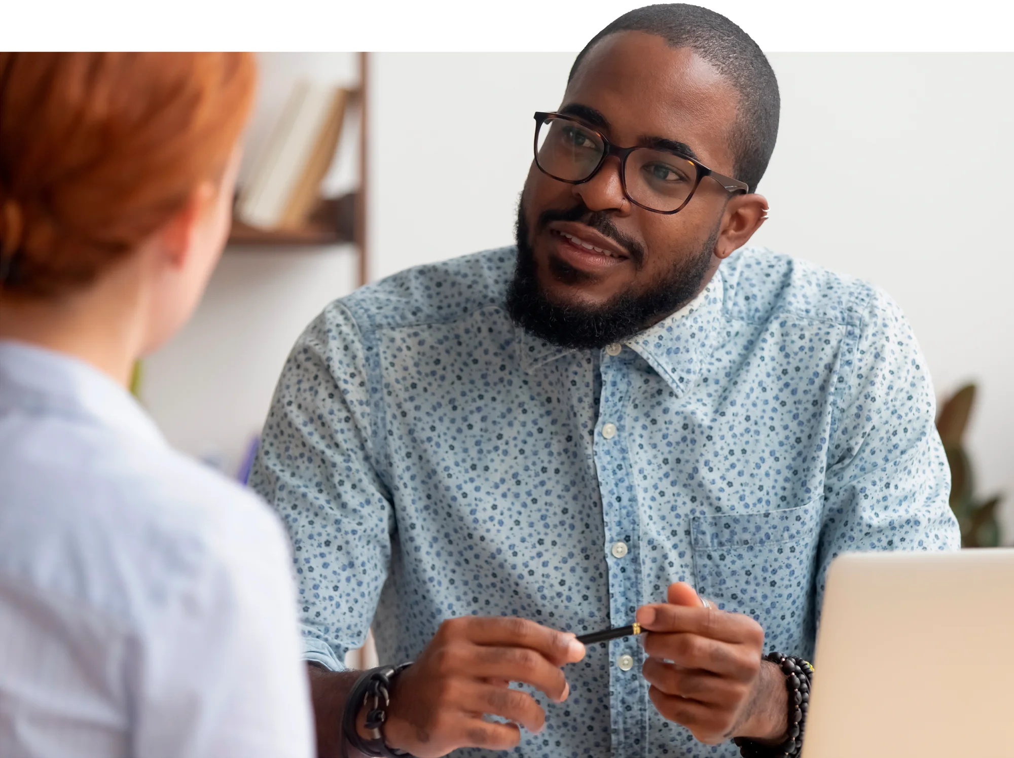 A man wearing glasses and a light blue shirt with a subtle pattern looks at a woman with red hair while sitting in a work environment.