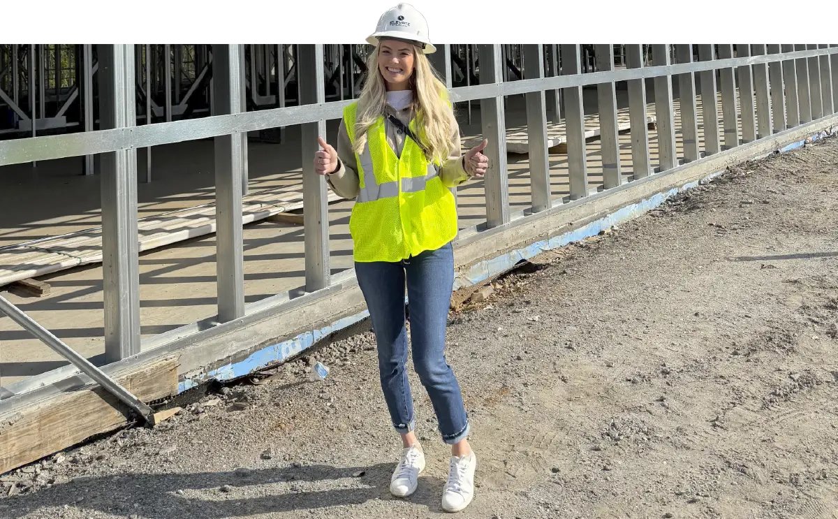 Courtney Cox wearing a hardhat and yellow safety vest while standing in front of a construction site with a smile and thumbs up. 