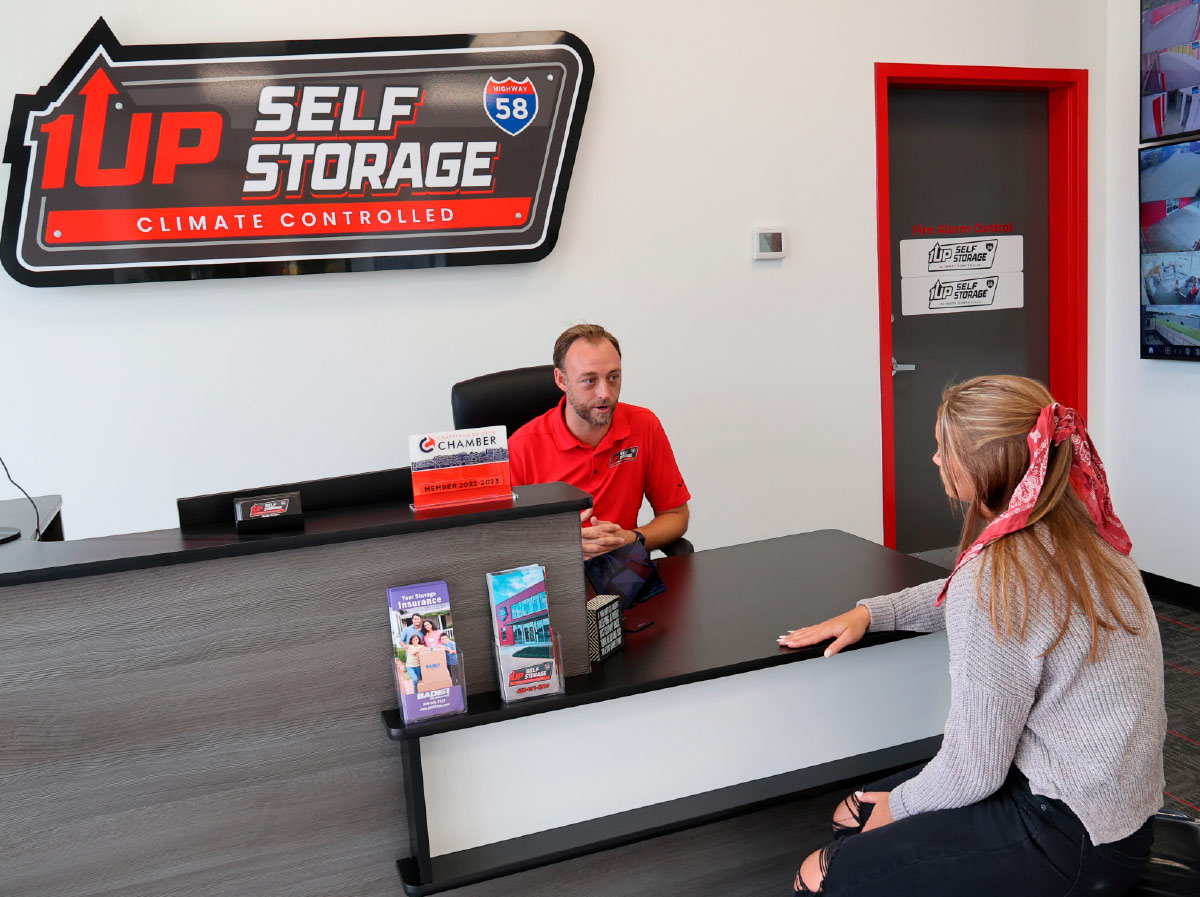 The front desk of 1up Self Storage facility. A male employee in a red shirt is seated behind a dark desk, speaking with a female customer.