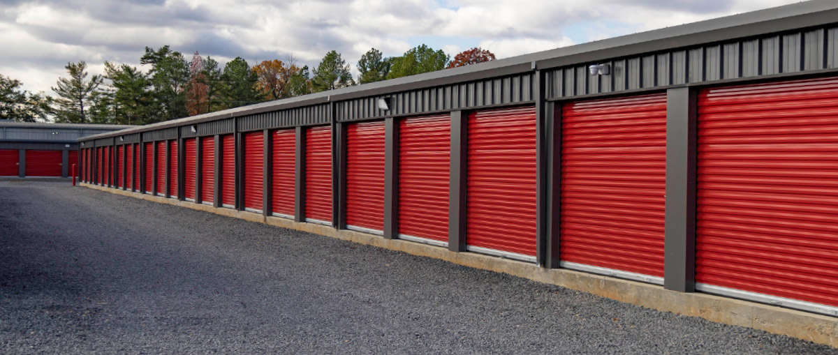 This image shows the exterior of a single-story self-storage building. The structure has dark gray, vertically-ribbed metal siding and a dark trim. The unit doors are a distinctive, vibrant red, corrugated roll-up metal, contrasting sharply with the gray building.