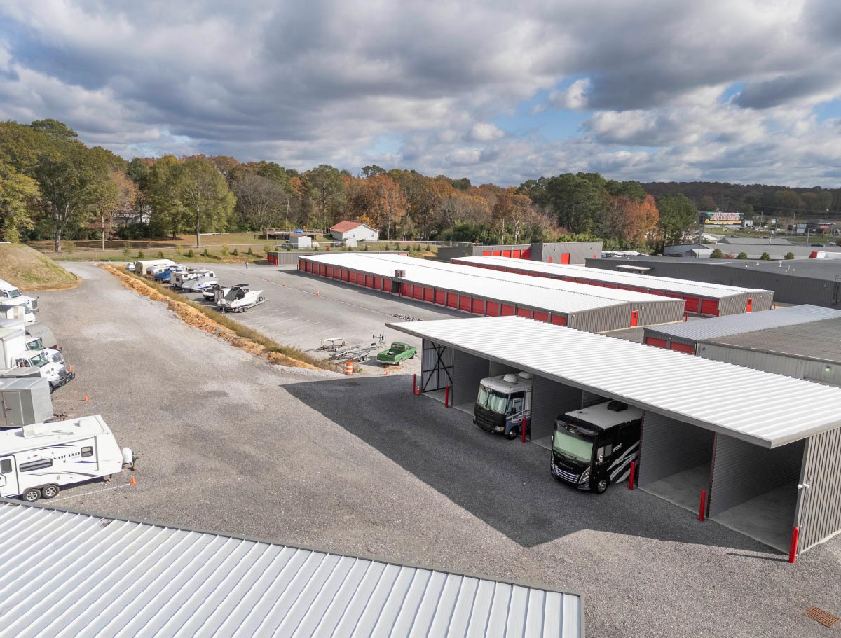 This is an aerial image of a large outdoor storage facility, which features covered parking structures shielding two large RVs/motorhomes, long rows of enclosed storage buildings with vibrant red roll-up doors, and an open gravel lot holding several trailers and boats.