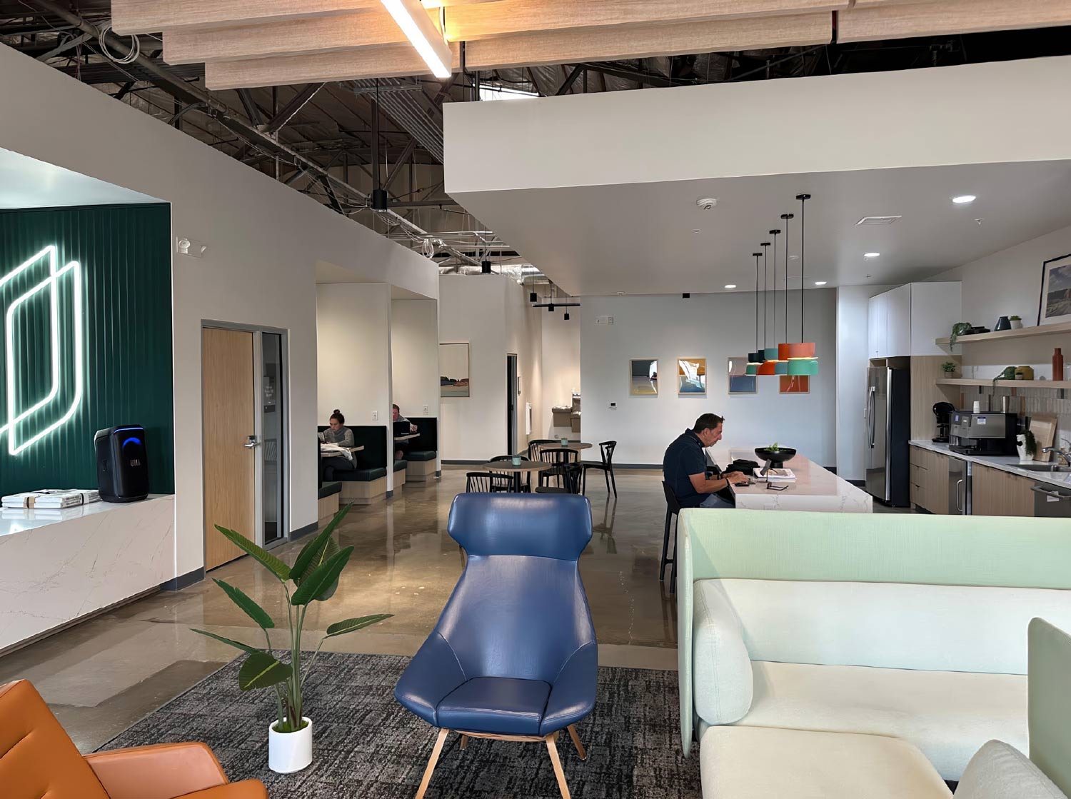 A bright, modern open-concept office space featuring a lounge and kitchen area. The foreground has a blue leather armchair and a cream-colored sofa on a gray rug. To the left, a dark green accent wall has a neon light sculpture. A man sits at a white kitchen island in the center, backed by light wood cabinetry. The space has high, exposed ceilings and polished concrete floors.