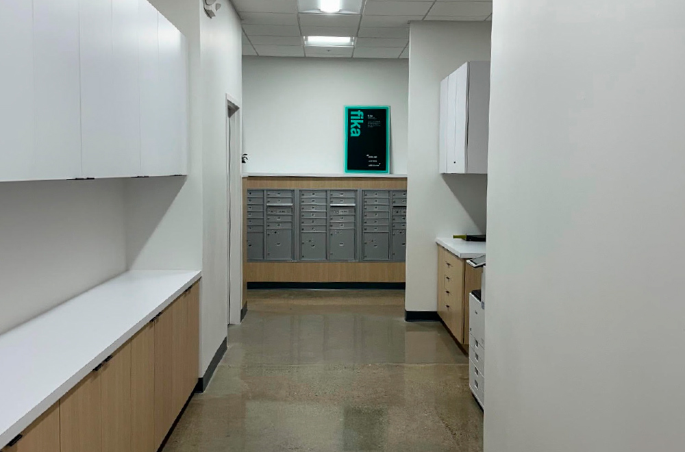 A view down a narrow, bright office hallway leading to a bank of gray mailboxes set into a light wood counter at the far end.
