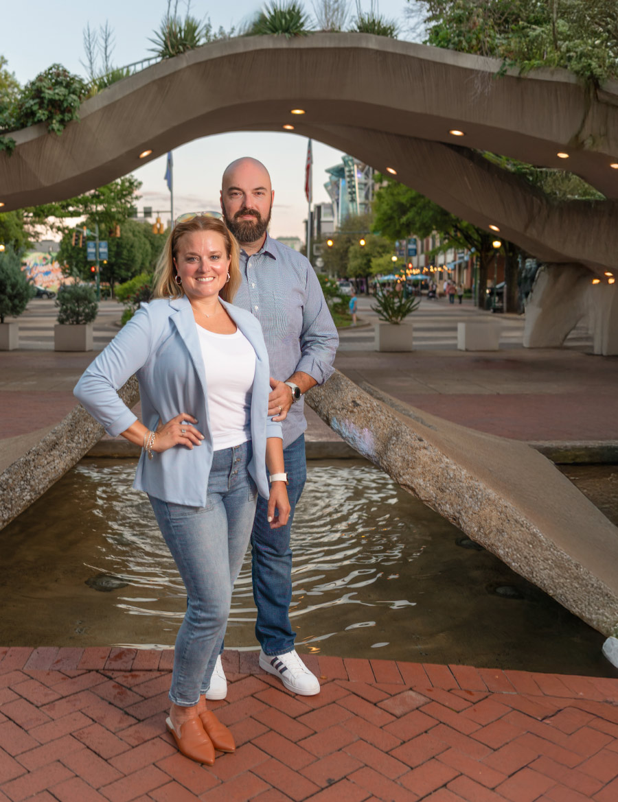 The Huffs standing one in front of the other while water feature and bridge is behind them