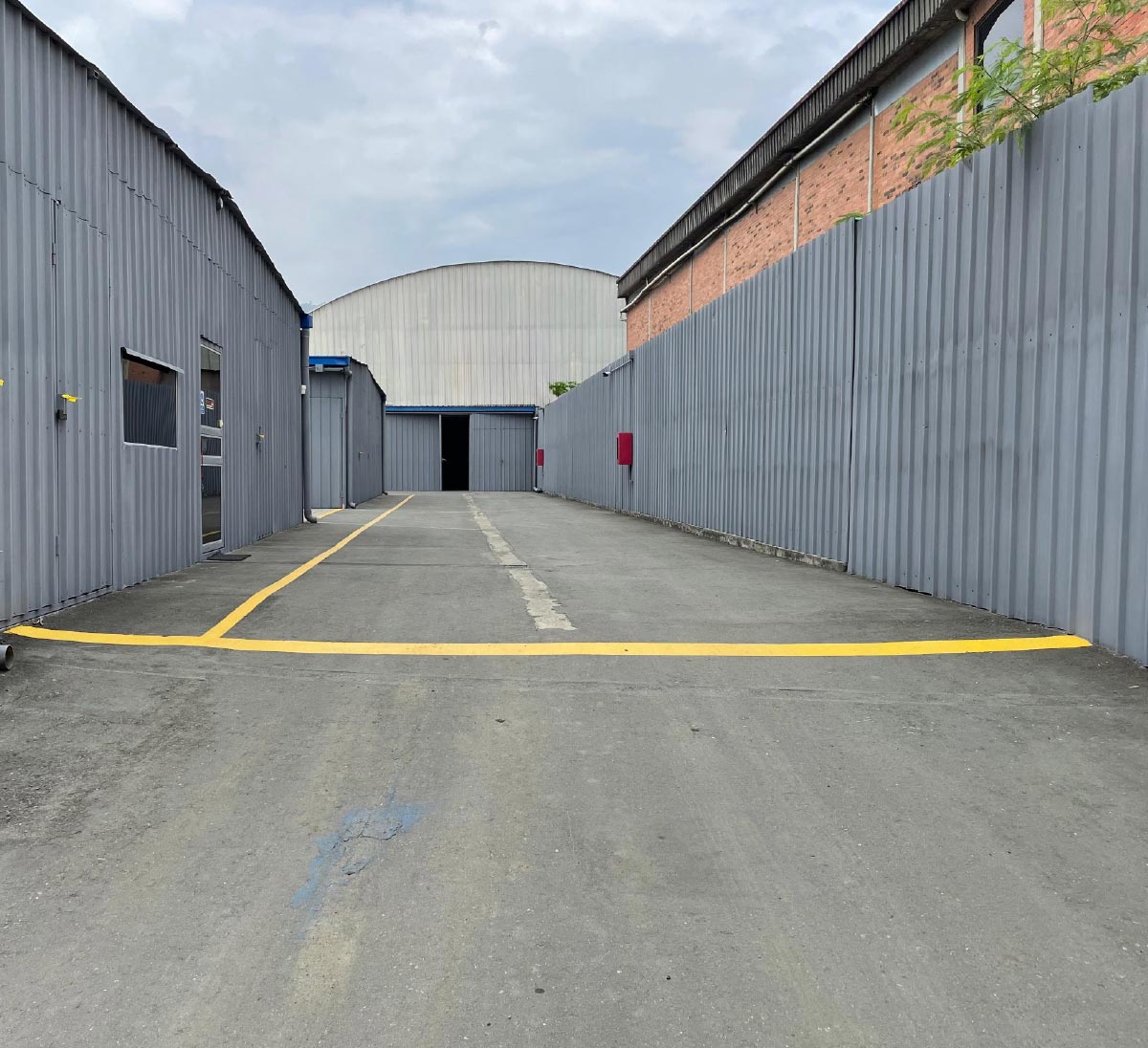 Paved industrial alley between gray corrugated metal walls. A yellow line is painted across the ground, leading to a large building ahead.