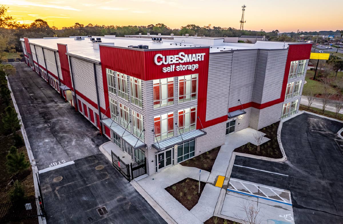  Aerial view of a large, modern CubeSmart Self Storage facility with red and gray paneling and an illuminated sign at sunset.