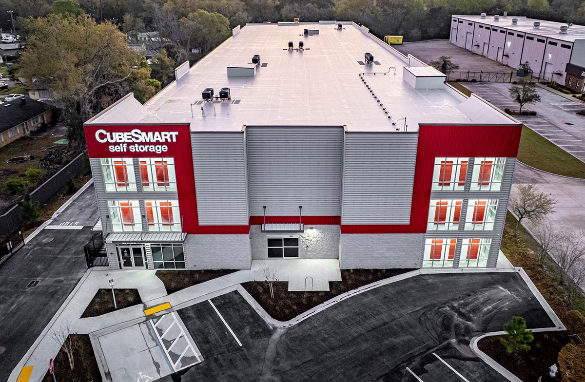  Aerial view of a large, modern CubeSmart Self Storage facility with a white roof, and red and gray exterior paneling at dusk.