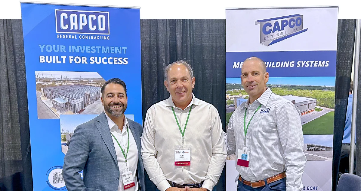 hree smiling Capco General Contracting representatives posing at a trade show booth with two large branded banners.