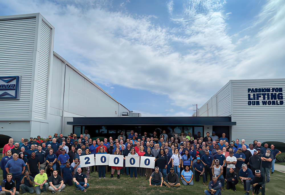 A large group of employees gathers outside a modern industrial building, holding signs to celebrate reaching a major milestone, "20,000".