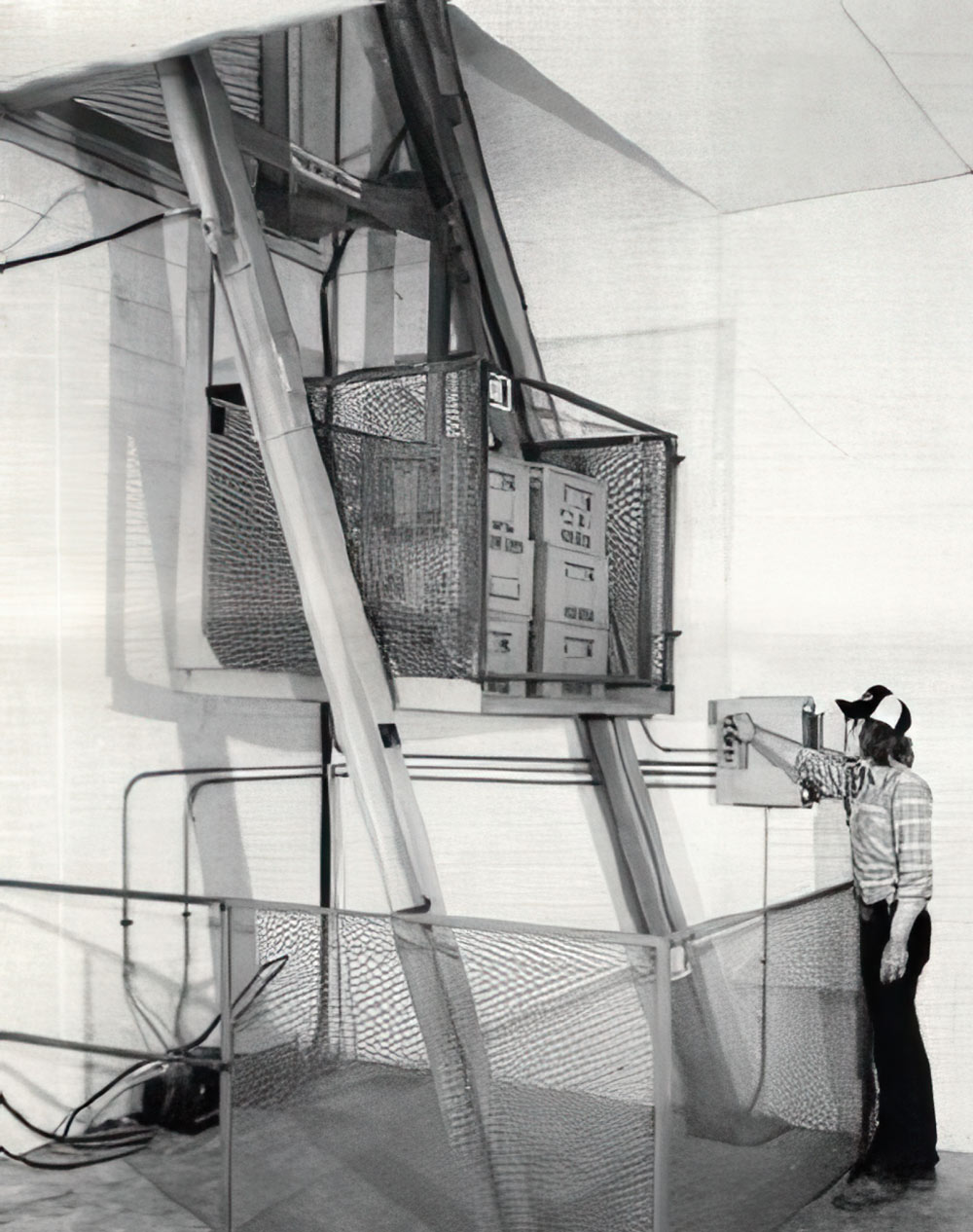 A black and white photo shows a woman operating the control panel of an early model vertical reciprocating conveyor (VRC), or freight lift.