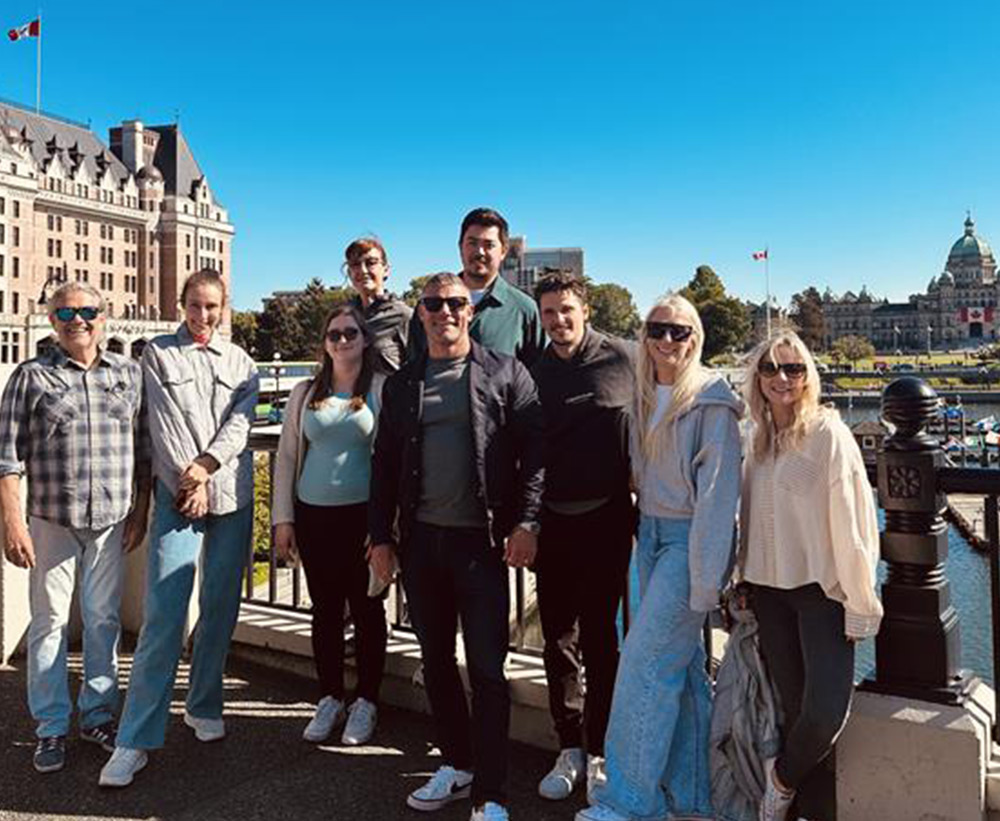 A group of smiling people posing outdoors on a sunny day in front of Victoria, BC landmarks.