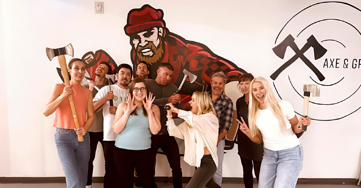 A group of adults smiling and posing with axes in an indoor axe-throwing venue with a lumberjack mural.