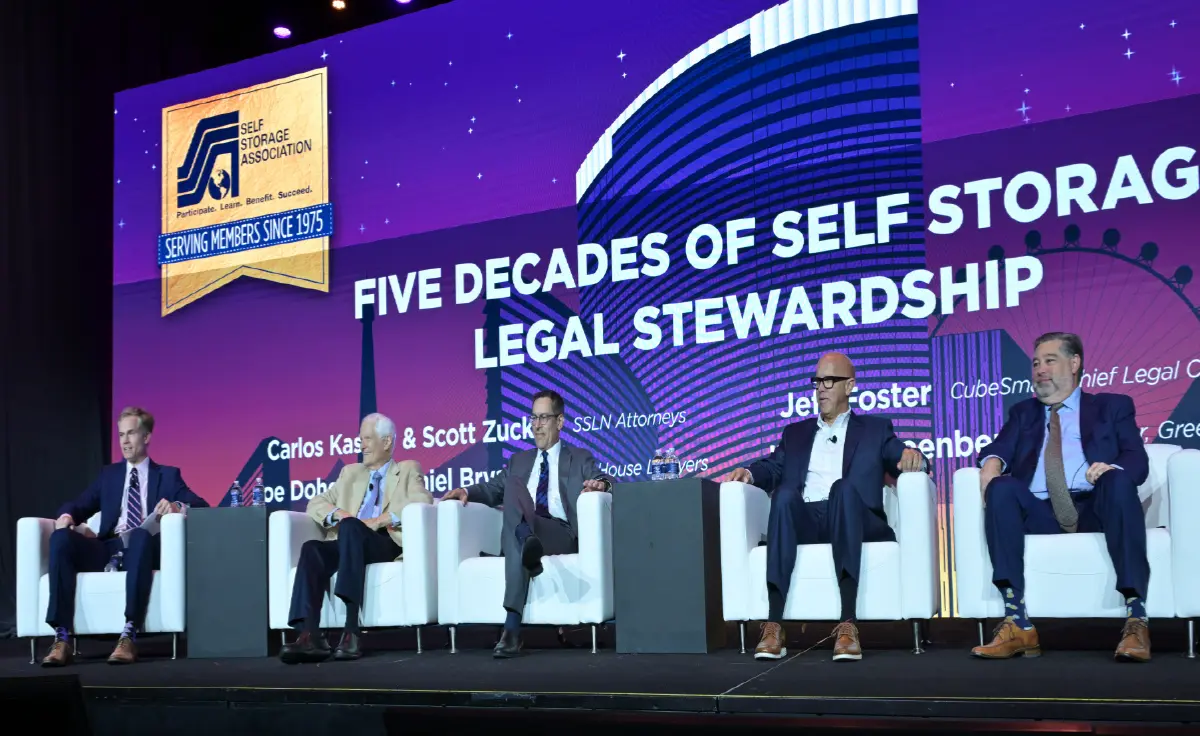 Five men in business attire are seated in white armchairs on a stage for a legal panel discussion with a large dominantly purple color/other multiple colors projector screen behind them; The projector screen displays the following - FIVE DECADES OF SELF STORAGE LEGAL STEWARDSHIP and the Self Storage Association logo; A few of the panelists' names, including Carlos Kaslow & Scott Zucker, Jeff Foster, etc., are partially visible on the projector screen