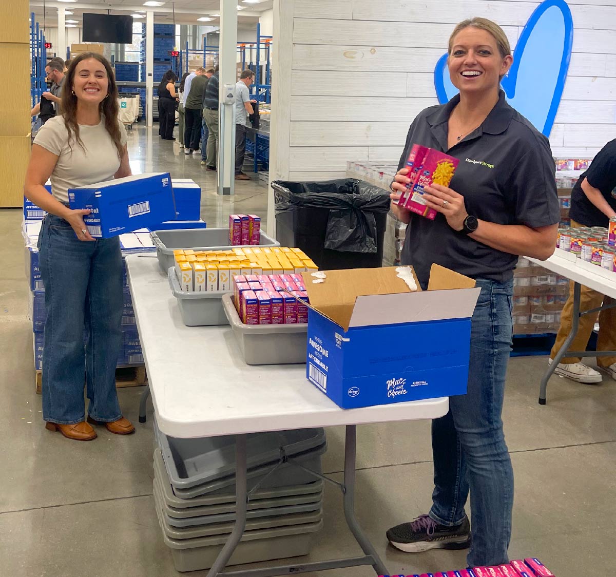 two women smiling while unpacking boxed goods and moving them to grey bins 