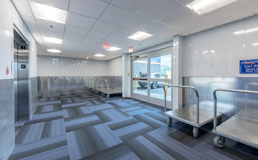 Clean indoor self-storage loading area with blue carpet, elevator, and several metal moving carts.
