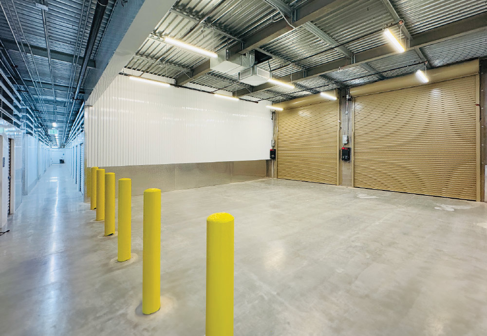Indoor loading bay with concrete floor, exposed ceiling, yellow bollards, and roll-up doors.