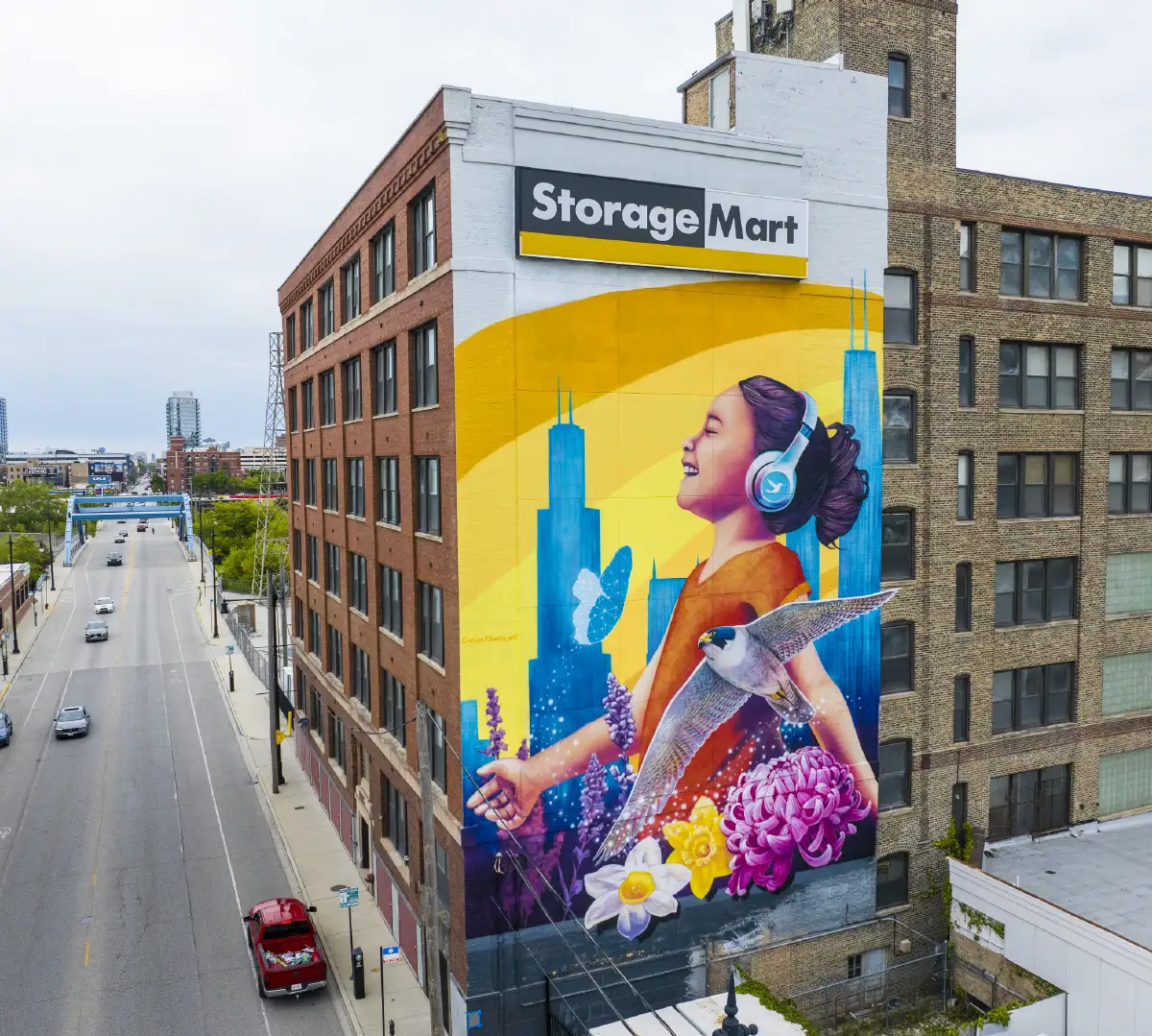 StorageMart facility with mural of a girl wearing headphones and smiling with flowers and a bird in the forefront and buildings in the background