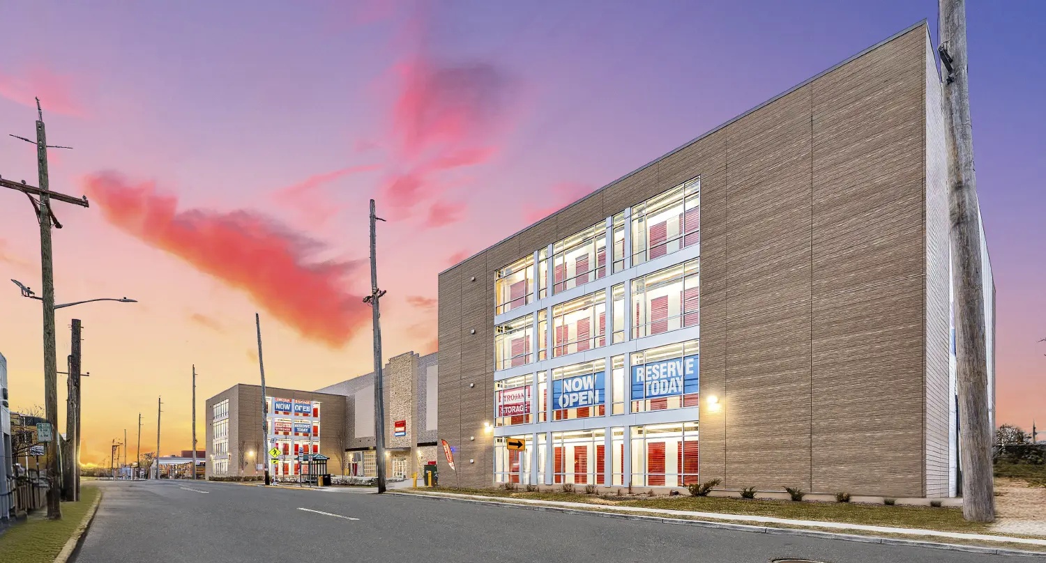 A wide exterior view of a large, multi-story self-storage facility with brown brick walls and rows of large windows, with "NOW OPEN" and "RESERVE TODAY" banners, photographed at dusk under a dramatic pink and purple sky.