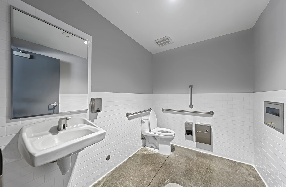 A wide-angle, ADA-compliant restroom in a commercial building with white subway tile walls, a gray-painted upper wall, a concrete floor, a wall-mounted sink, and a toilet with grab bars.