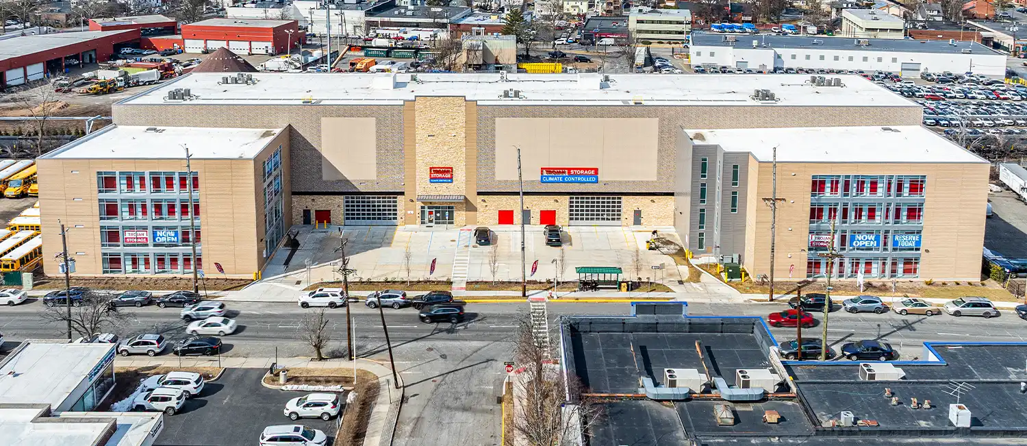 An overhead view of a large, multi-story, modern climate-controlled self-storage facility with beige facade panels, red roll-up doors, and red window treatments, surrounded by an urban setting with multiple cars in the parking lot.