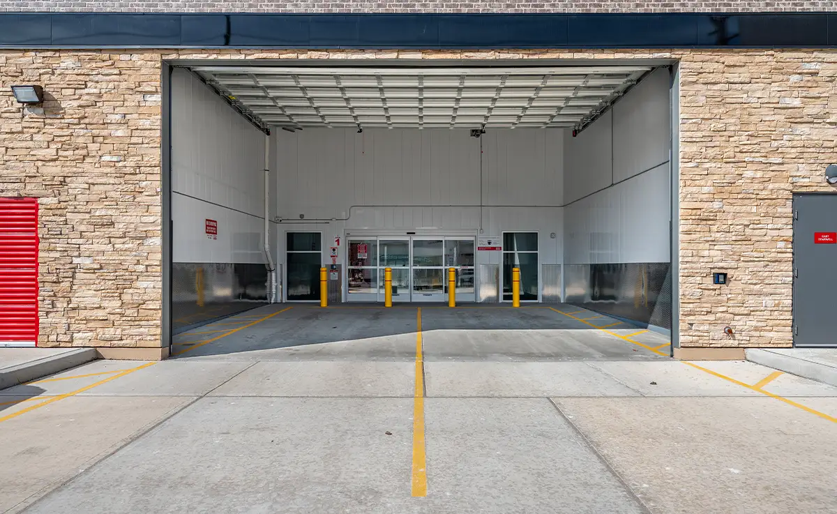 The drive-in entry bay of a self-storage facility features a high, open ceiling, light stone veneer side walls, and a glass automatic sliding door entrance flanked by yellow bollards and marked concrete paving.